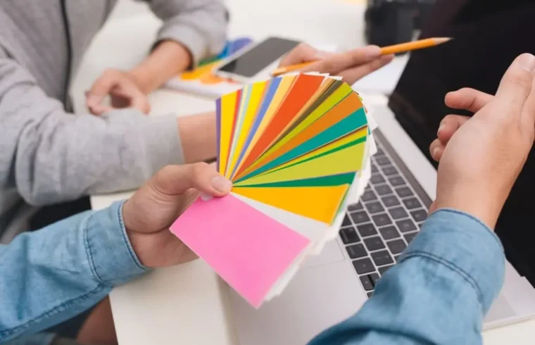 Mãos segurando cores em frente a um computado para escolher a melhor paleta de cores para o seu projeto digital