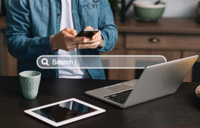 homem-de-camisa-jeans-e-branca-usando-celular-notebook-e-tablet-pesquisa-sobre-otimizacao-de-ecommerce-com-uma-barra-de-busca-no-centro-da-imagem